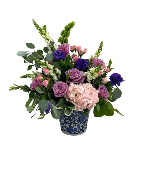 An arrangement of pink hydrangeas, lavender roses, purple lisiathus, white snapdragons and pink spray roses in a blue and white ceramic vase